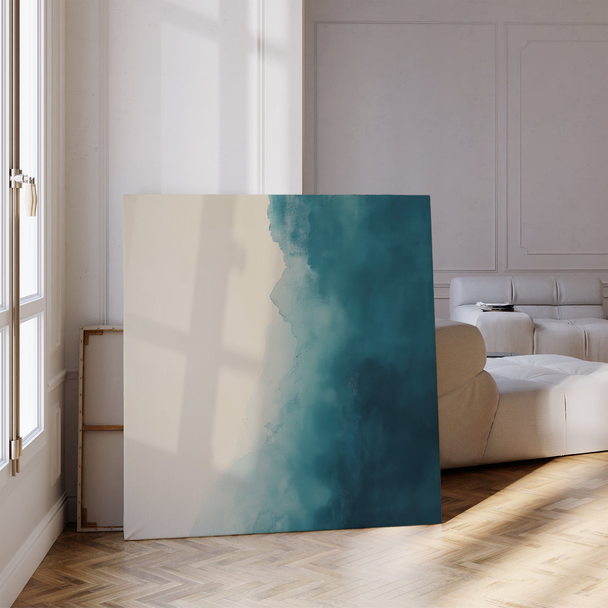 The Teal Tide Canvas Art, featuring cream and deep blue tones, leans against the wall of a bright living room with wooden floors, a beige sofa, and sunlight pouring through tall windows.