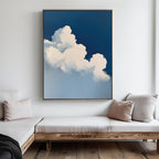 A modern living room with light wood furniture and neutral cushions, highlighted by calming decor and The Cumulus Abstract Canvas Art—a large framed painting of fluffy white clouds on a deep blue sky—adding minimalist style to the white wall.