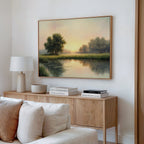 A cozy living room features a light wood sideboard with stacked books, a lamp, and decor. Above it hangs The Journey Canvas Art—a rustic painting of a calm river at sunrise or sunset, adding countryside charm to the space.