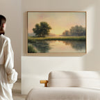 A person stands in a softly lit room, admiring The Journey Canvas Art—a framed lakeside sunrise scene with trees—hanging above a beige sofa, bringing rustic countryside decor charm to the tranquil space.