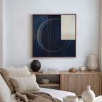 A modern living room features a beige sofa, neutral pillows, a wooden sideboard with vases and books, and The Orbit Canvas Art—an abstract blue and cream circle design—on a white wall. Warm natural light flows in, complementing the stylish decor.