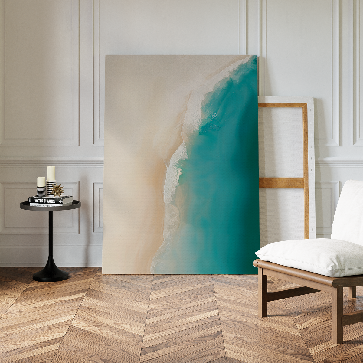 The Beach Abstract Canvas Art, a large coastal wall piece with a turquoise aerial beach scene, leans against a white-paneled wall. A black table with candles and books and a wooden chair rest on the herringbone floor nearby.