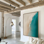 A modern living room with exposed wooden beams, white walls, a curved floor lamp, and The Beach Abstract Canvas Art—a large framed piece showing an aerial view of turquoise sea meeting sandy shore.