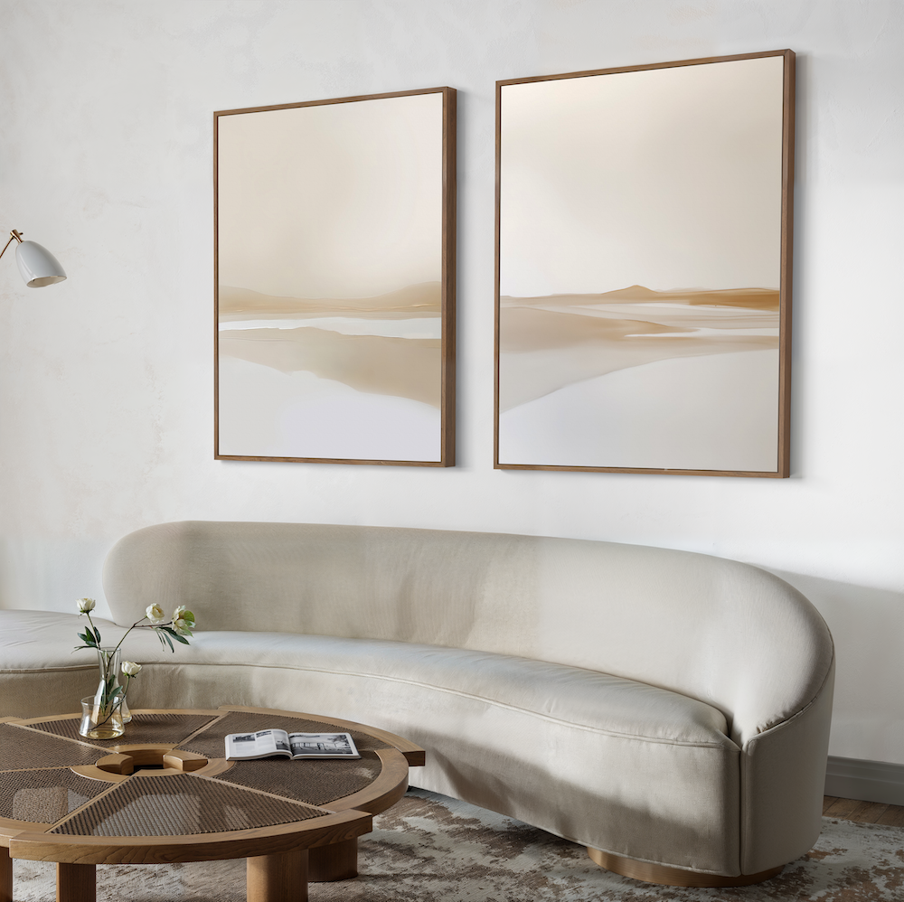 A modern living room features a curved beige sofa, a round wooden coffee table, a small vase with flowers, and two Tranquil Horizons Set of 2 abstract landscape paintings in neutral tones on a white wall for an inviting touch.