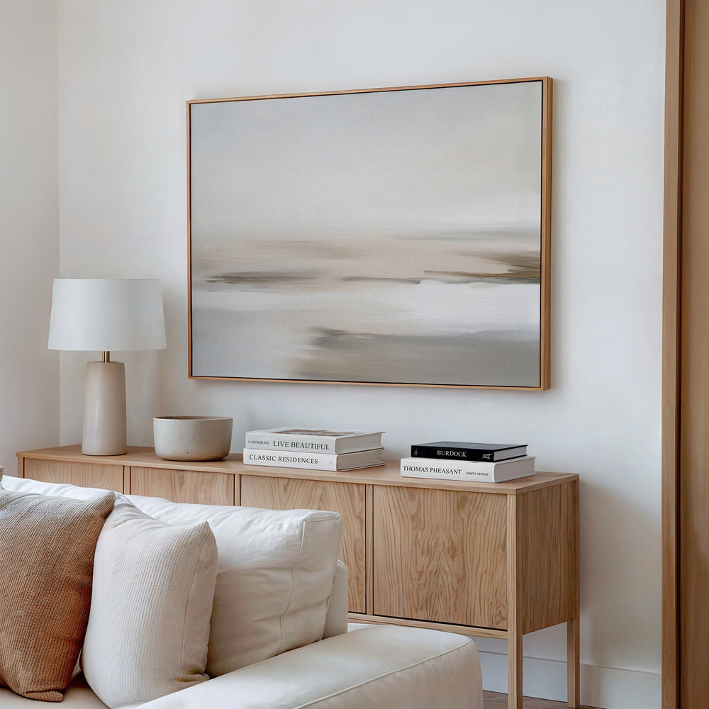 A minimalist living room features a wooden sideboard, white table lamp, stacked books, bowl, beige couch, and the Tranquil Shifts Canvas Art – Horizontal in neutral tones on a white wall for an elegant decor finish.