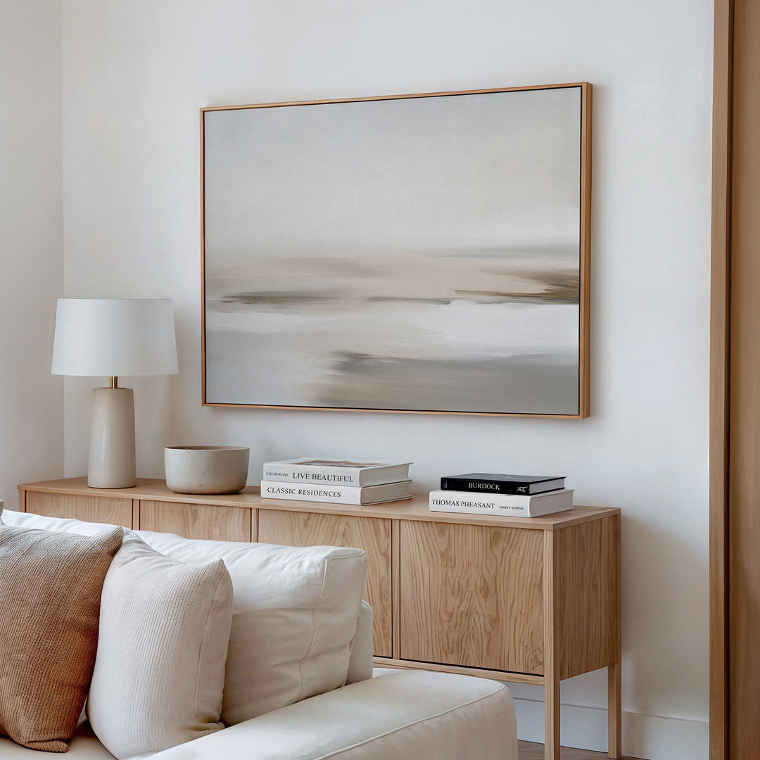 A minimalist living room features a wooden sideboard, white table lamp, stacked books, bowl, beige couch, and the Tranquil Shifts Canvas Art – Horizontal in neutral tones on a white wall for an elegant decor finish.
