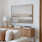 A minimalist living room features a wooden sideboard, white table lamp, stacked books, bowl, beige couch, and the Tranquil Shifts Canvas Art – Horizontal in neutral tones on a white wall for an elegant decor finish.