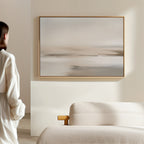 A person in a white robe stands by a beige couch, admiring the Tranquil Shifts Canvas Art – Horizontal in neutral tones on a cream wall, enhancing the minimalist living room decor.