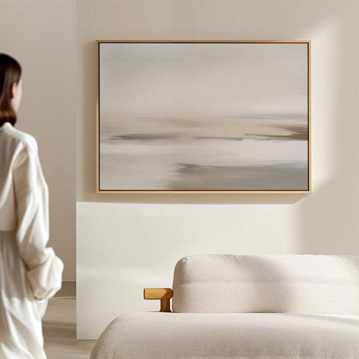 A person in a white robe stands by a beige couch, admiring the Tranquil Shifts Canvas Art – Horizontal in neutral tones on a cream wall, enhancing the minimalist living room decor.