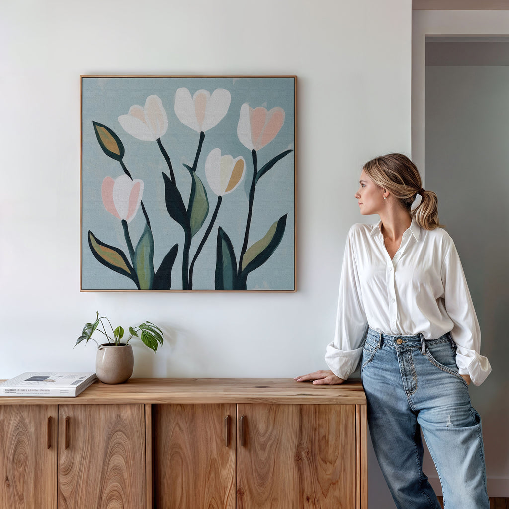 A woman in a white blouse and jeans stands by a wooden cabinet, admiring the Tulip Harmony Canvas Art—minimalist wall decor with pastel flowers. Perfect for living rooms, it’s accented by a potted plant and books on the cabinet below.