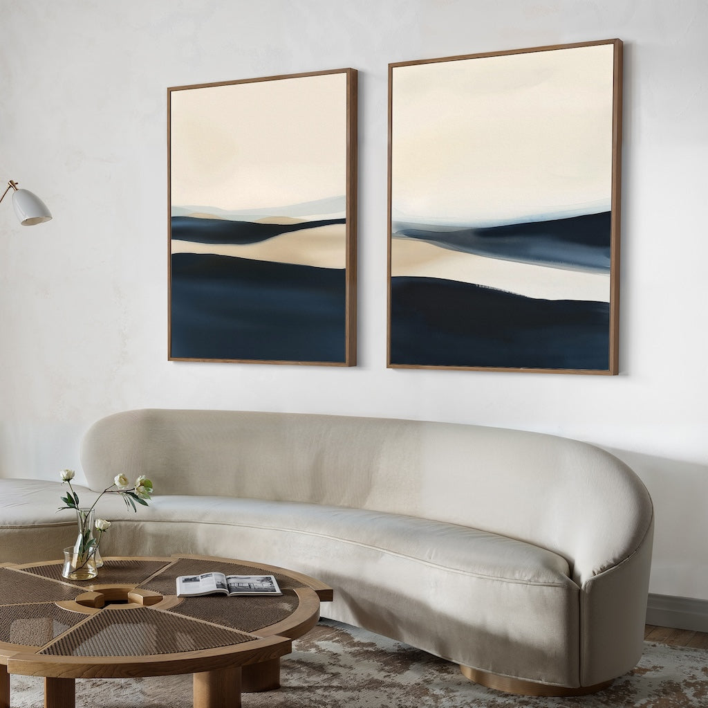A modern living room with a curved beige sofa, a round wood-and-glass coffee table, and the Twilight Horizons Set of 2 minimalist abstract landscape paintings in neutral and dark tones creates a welcoming contemporary atmosphere.