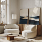 A modern living room features the Twilight Horizons Set of 2 abstract paintings in blue and beige, paired with two rounded white armchairs, a low wooden coffee table, and minimalist decor. Sunlight through large windows enhances the serene ambiance.