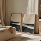 The Twilight Horizons Set of 2 abstract paintings in blue, beige, and cream tones lean against a softly lit wall beside a cozy cream armchair and wooden cabinet—ideal accents for modern home decor.