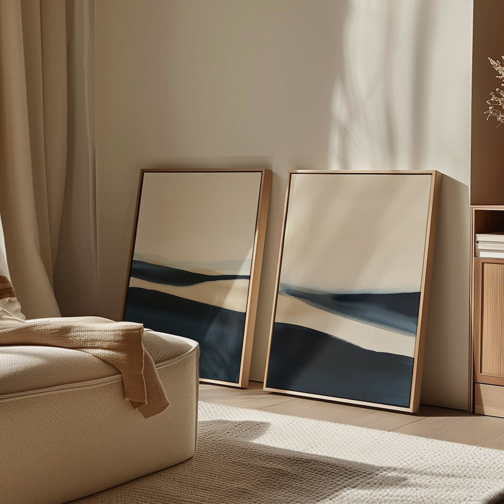 The Twilight Horizons Set of 2 abstract paintings in blue, beige, and cream tones lean against a softly lit wall beside a cozy cream armchair and wooden cabinet—ideal accents for modern home decor.