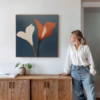 A woman in a white shirt and jeans stands by a wooden cabinet, gazing at the Twin Blooms Canvas Art of two abstract flowers above her. A small plant and book on the cabinet complete the Nordic Park-inspired decor.