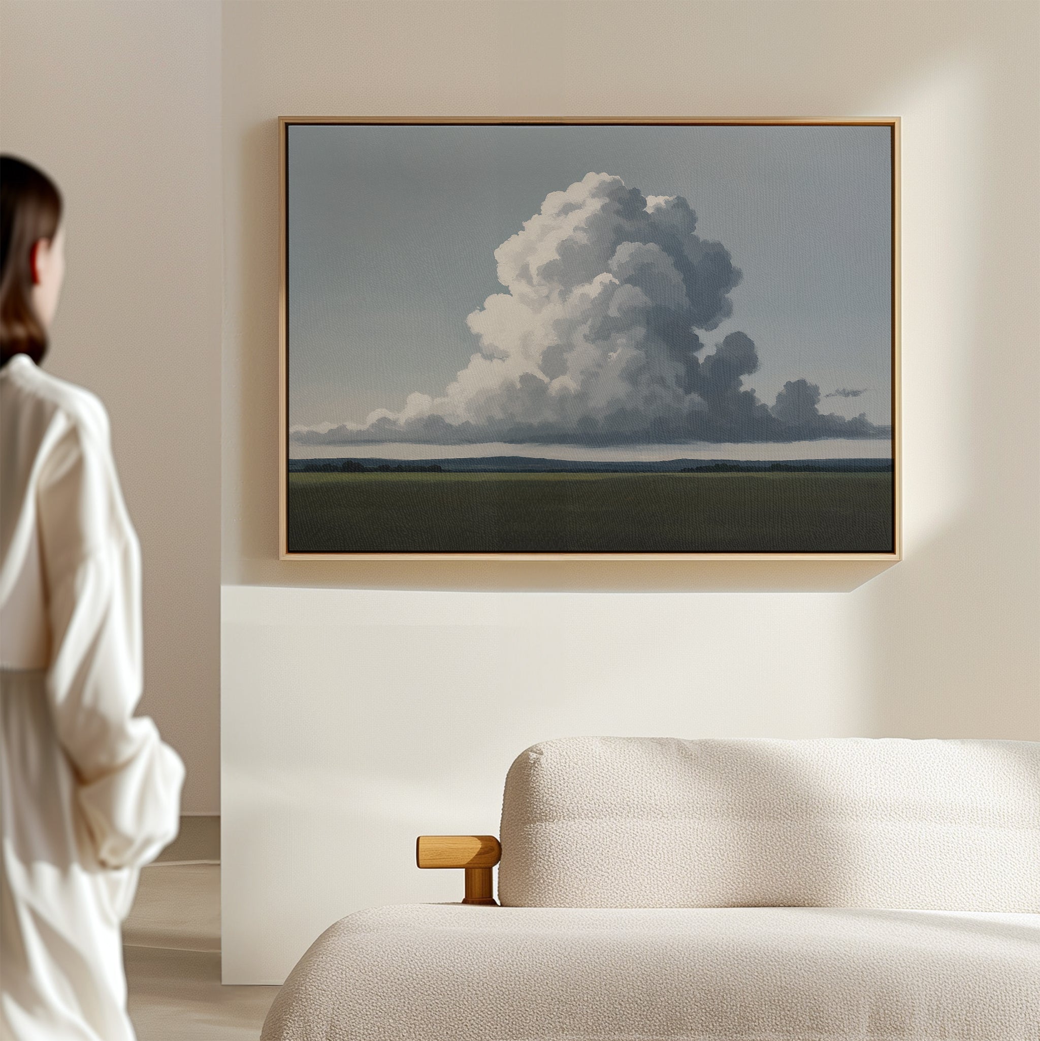 A person in white admires the Uplifted Sky Canvas Art—a minimalist piece depicting a large cloud over a flat landscape—perfect for enhancing modern bedrooms with wall art above a beige sofa.