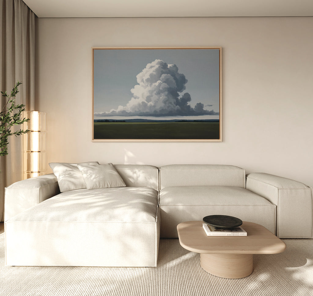 A modern living room with a beige sectional, light wood coffee table, potted plant, and Uplifted Sky Canvas Art depicting clouds and a green field. Neutral tones and soft lighting create a calm, serene atmosphere.