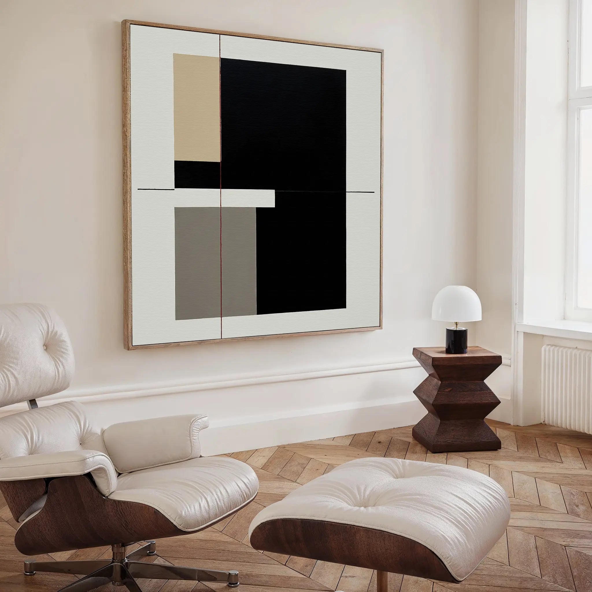 A modern, minimalist living space featuring a cream Eames lounge chair and ottoman, a geometric wooden side table with a small white lamp, and Urban Balance Canvas Art in black, beige, and gray blocks displayed on the wall.