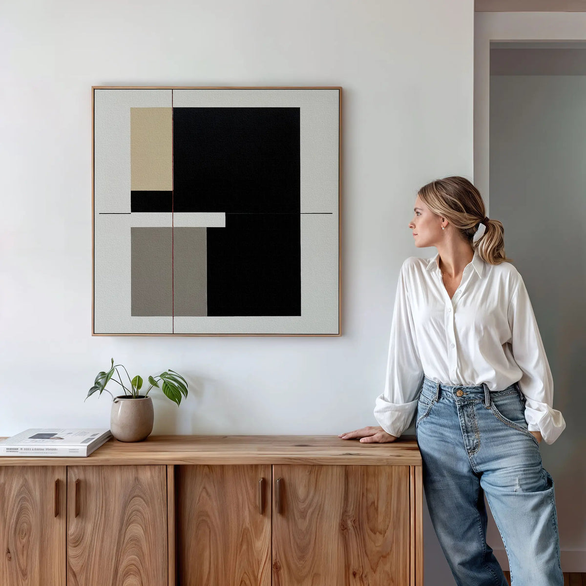 A woman in a white shirt and jeans leans on a wooden sideboard with a plant and book, gazing at the Urban Balance Canvas Art—minimalist wall decor with black, beige, and gray shapes—mounted on a white wall.