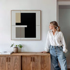 A woman in a white shirt and jeans leans on a wooden sideboard with a plant and book, gazing at the Urban Balance Canvas Art—minimalist wall decor with black, beige, and gray shapes—mounted on a white wall.