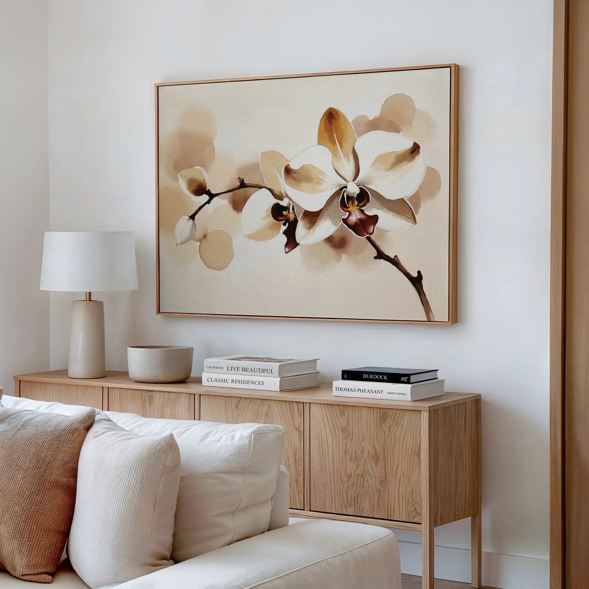 A modern living room with a beige couch, light wood sideboard, stacked books, lamp, and elegant wall decor—a large Vanilla Bloom Canvas Art featuring white orchids with brown accents enhances the space.