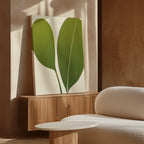 The Verdant Duo Abstract Canvas Art, depicting two large green leaves, leans against a wooden cabinet in a softly lit modern room with neutral tones, stylish wall art, a round white table, and a textured white sofa.