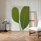 The Verdant Duo Abstract Canvas Art, featuring two green leaf shapes, leans against a white paneled wall in a modern living room beside a black side table with candles and books and a white cushioned chair on herringbone wood floors.