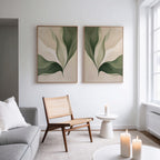A stylish living room features a woven chair, small white table, candles, gray sofa, and two minimalist Verdant Grace Set of 2 wall art pieces with abstract green leaves. Natural light from a tall window enhances the decor.