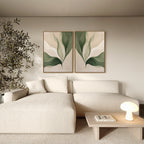 A modern living room with a cream textured sectional sofa, Verdant Grace Set of 2 minimalist wall art featuring green and beige leaf prints, a small wooden coffee table with a round lamp, and a plant in the corner.