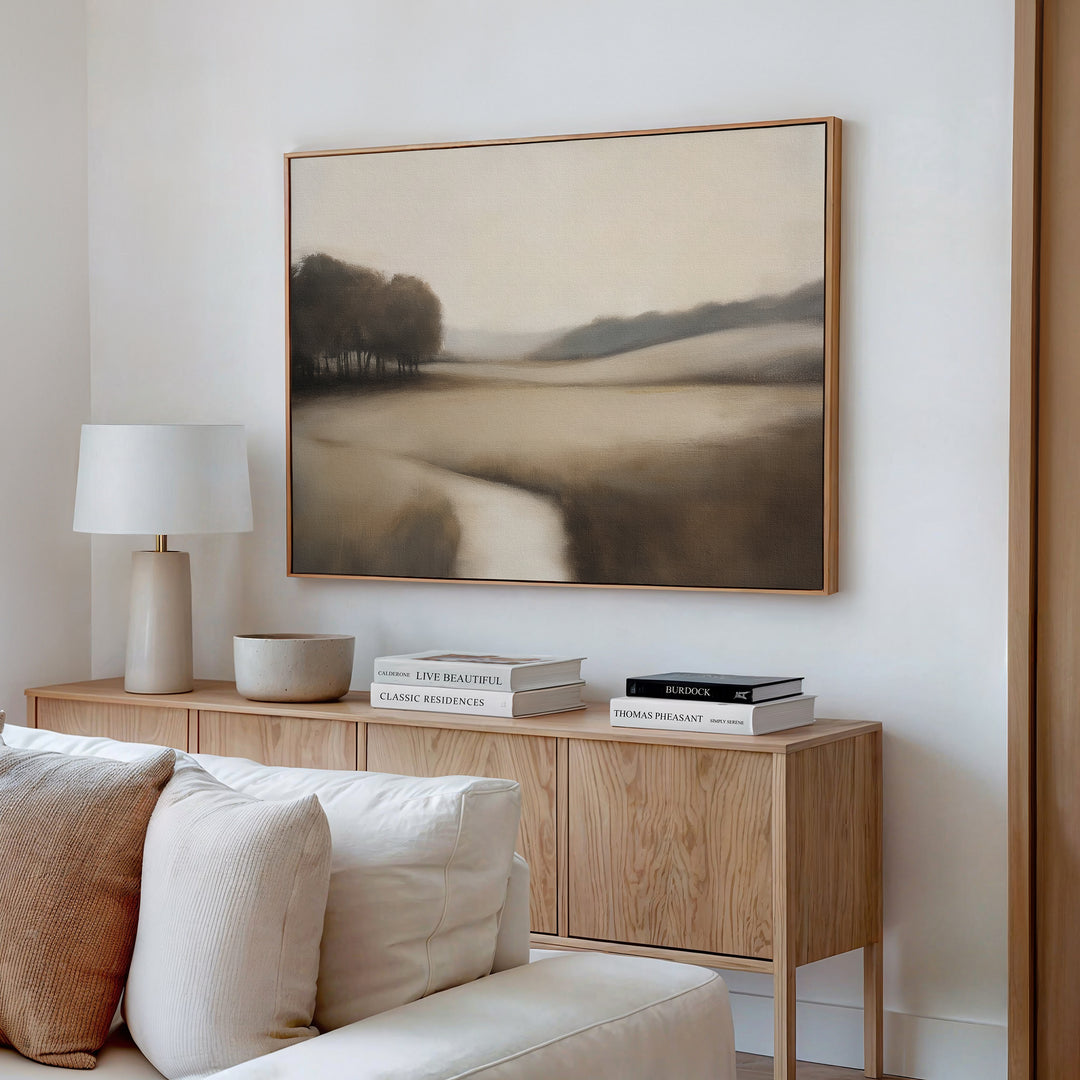 A modern living room features a beige sofa, a wooden sideboard with books, a lamp, and a bowl, and the Vintage Horizon Canvas Art – Horizontal displaying serene trees and river in neutral tones on the wall.