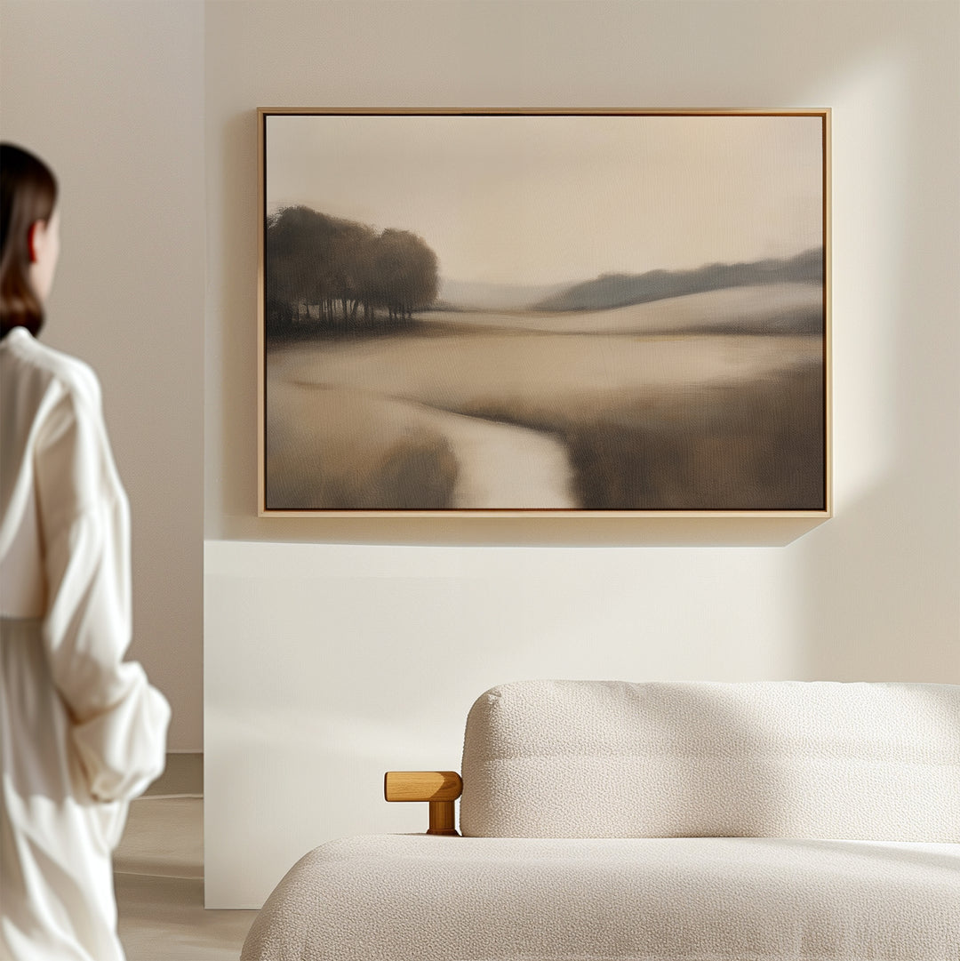 In a minimal, light-filled room, a person admires Vintage Horizon Canvas Art – Horizontal: a serene landscape print with soft, muted tones and silhouettes of trees beside a winding river on a textured sofa.