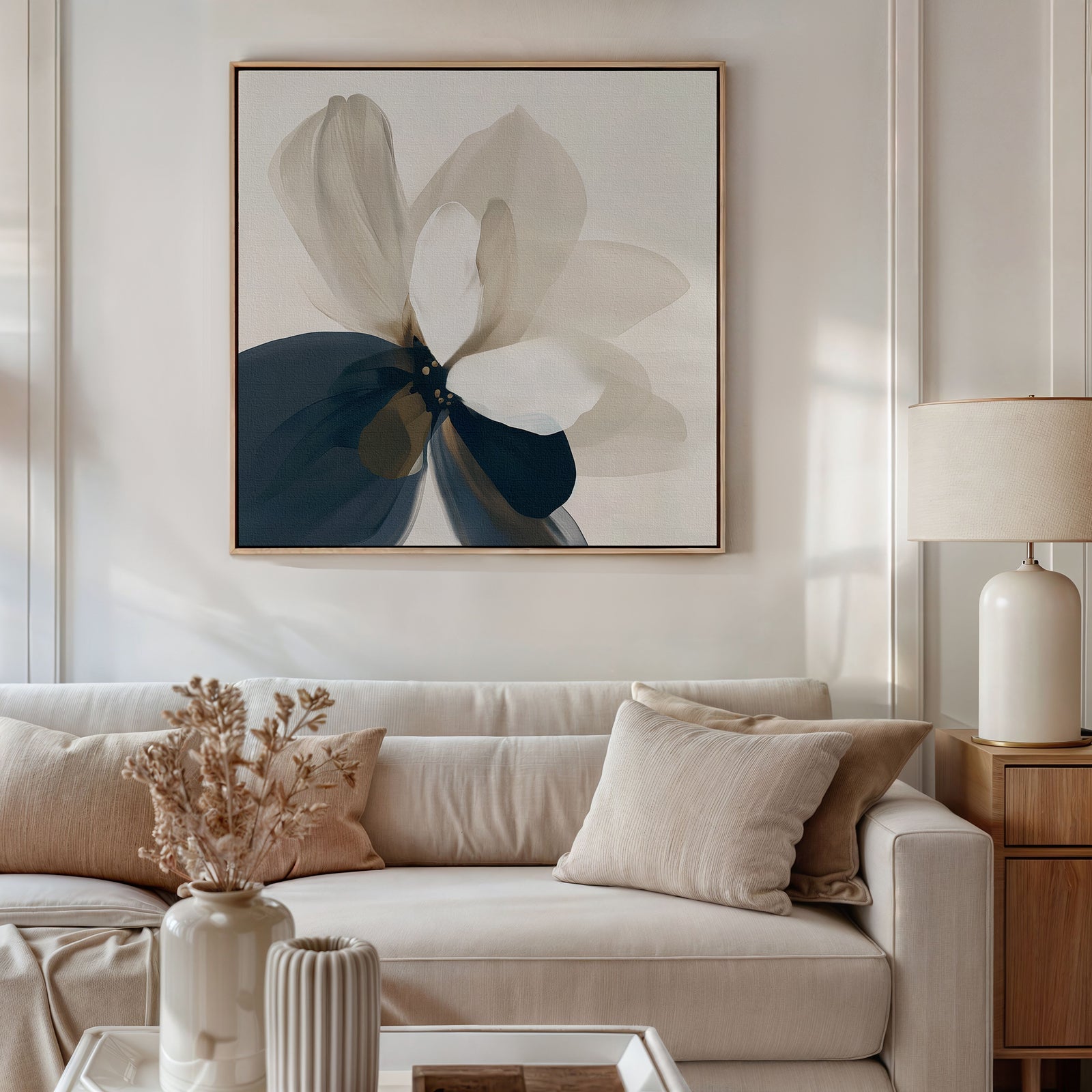 A modern living room with a beige sofa, neutral cushions, and dried flowers in a vase on the coffee table. A large abstract flower painting hangs above the sofa, and a ceramic lamp sits on a wooden side table.