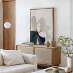 A modern living room with neutral tones featuring Whispering Petals Canvas Art - Vertical, a light wood sideboard, potted plant, decorative vases, stacked books, and a white sofa with cream cushions—perfect for stylish decor.