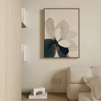 A minimalist living room corner features a beige sofa, the Whispering Petals Canvas Art - Vertical with an abstract flower design, and shelves with white books and a small decorative object for a serene, neutral atmosphere.