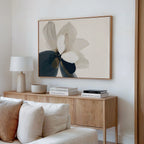 A minimalist living room with a light wooden sideboard, neutral decor, a white table lamp, stacked books, a ceramic bowl, and the Whispering Petals Canvas Art - Horizontal displayed above a cream-colored sofa.