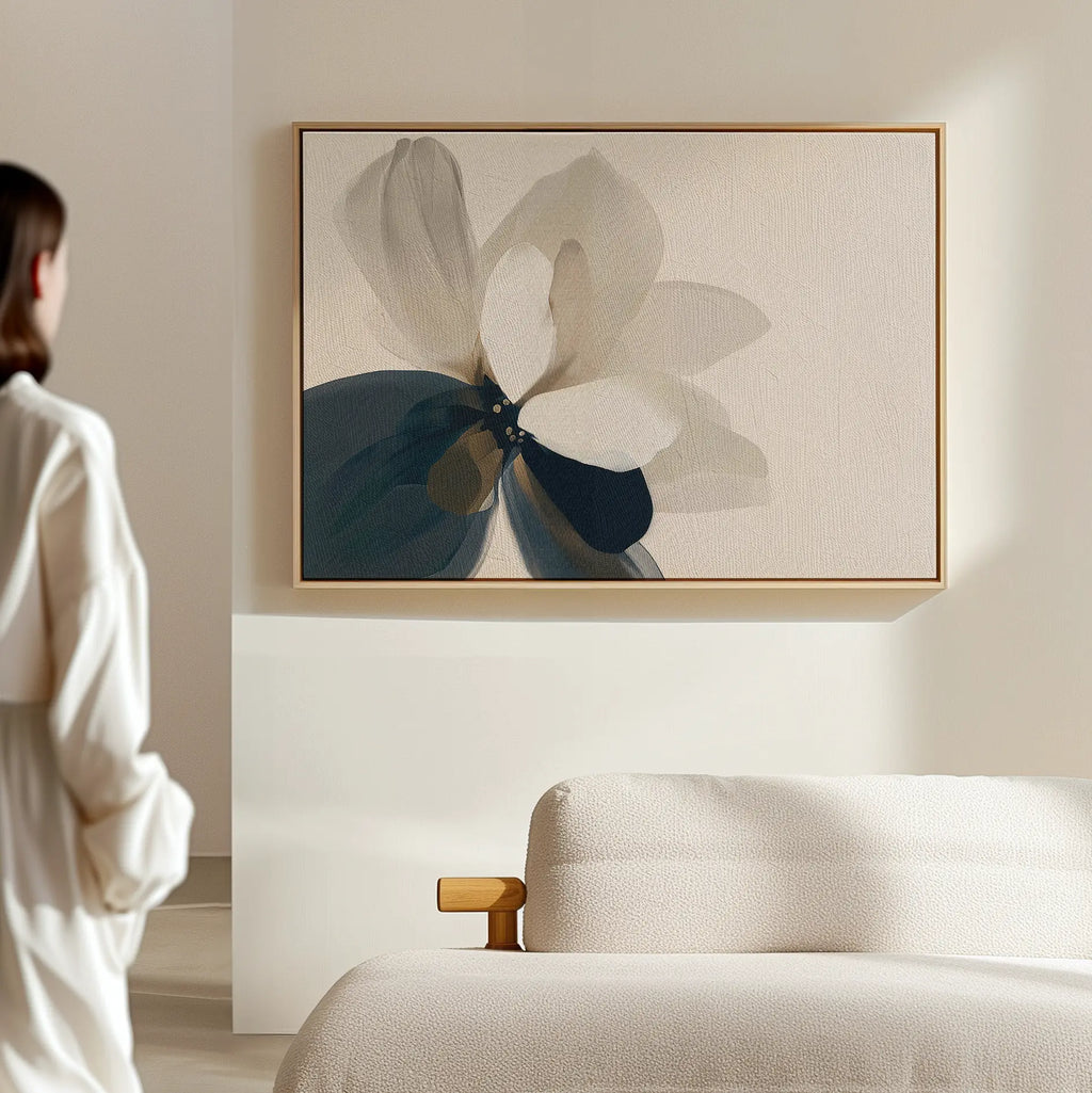 A woman in a white outfit admires the Whispering Petals Canvas Art - Horizontal, a large abstract flower in beige, blue, and gray tones, hanging above a cream-colored bed as elegant bedroom decor in a minimal, light-filled room.