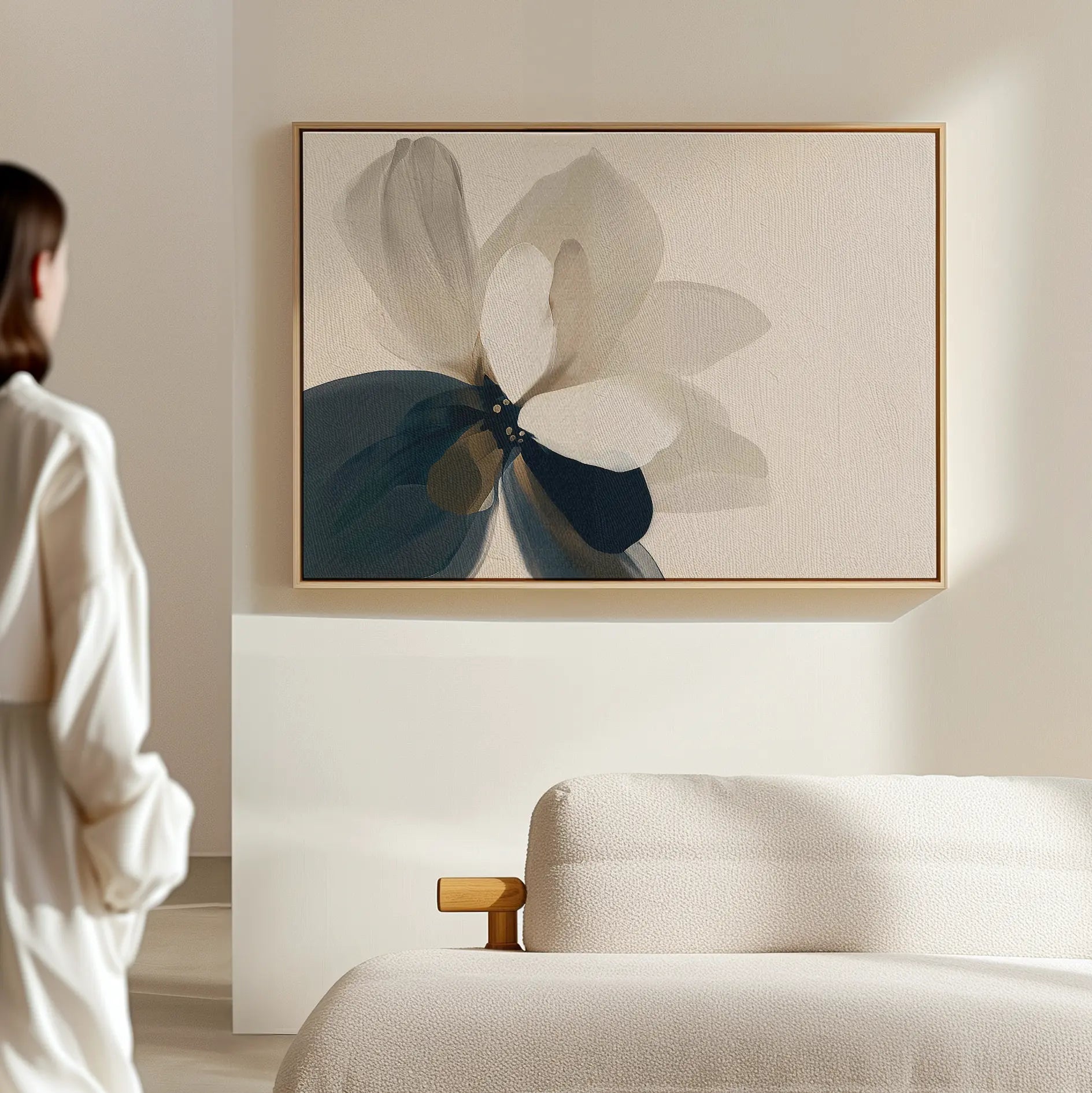 A woman in a white outfit admires the Whispering Petals Canvas Art - Horizontal, a large abstract flower in beige, blue, and gray tones, hanging above a cream-colored bed as elegant bedroom decor in a minimal, light-filled room.