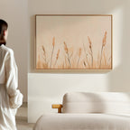 A person in white admires the Whispering Reeds Canvas Art, which features soft beige grasses, displayed above a light-textured sofa in a modern, bright room.