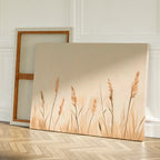 Two canvases rest on a wooden floor against a white wall; one is turned to show its frame while the other displays Whispering Reeds Canvas Art, featuring beige and brown wheat-like plants—ideal minimalist wall art for your living room decor.