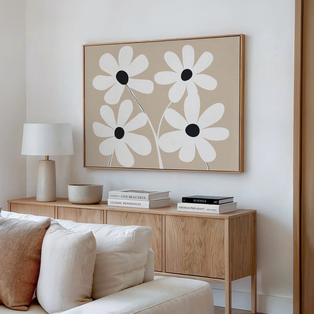 A modern living room features the White Blooms Canvas Art, a light wood sideboard, white lamp, stacked books, ceramic bowl, neutral-toned decor, and a cream sofa. The wall art shows four white flowers with black centers.