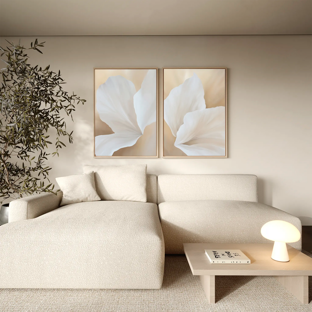 A modern living room with a light beige sofa, wooden coffee table with a lamp and books, potted plant, and the White Flora Set of 2 botanical wall art featuring large white flowers above the sofa.