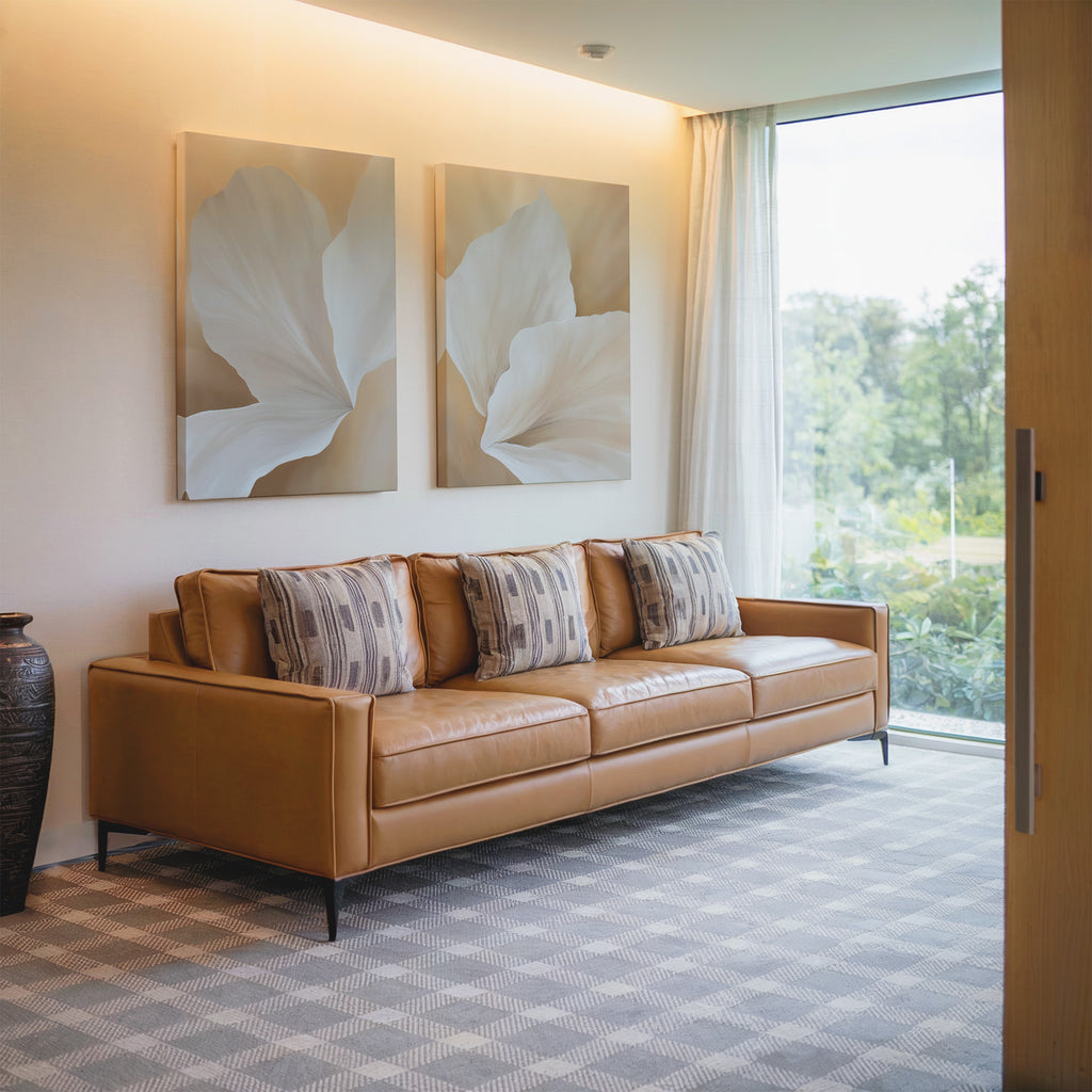 Modern living room with a tan leather sofa, patterned cushions, floor-to-ceiling windows overlooking Nordic Park greenery, and two White Flora Set of 2 botanical wall art pieces featuring white flora above a tall side vase.