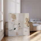 The White Whispers Canvas Art, featuring abstract floral designs in soft beige and white, leans against the wall of a bright living room with herringbone floors and a cream sofa, as sunlight enhances its minimalist charm.