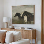 The Wild Current Canvas Art, a minimalist piece with three running horses, hangs above a wooden sideboard holding books, a lamp, a bowl, and a vase. A cream sofa adds to the neutral tones of this stylish living room.