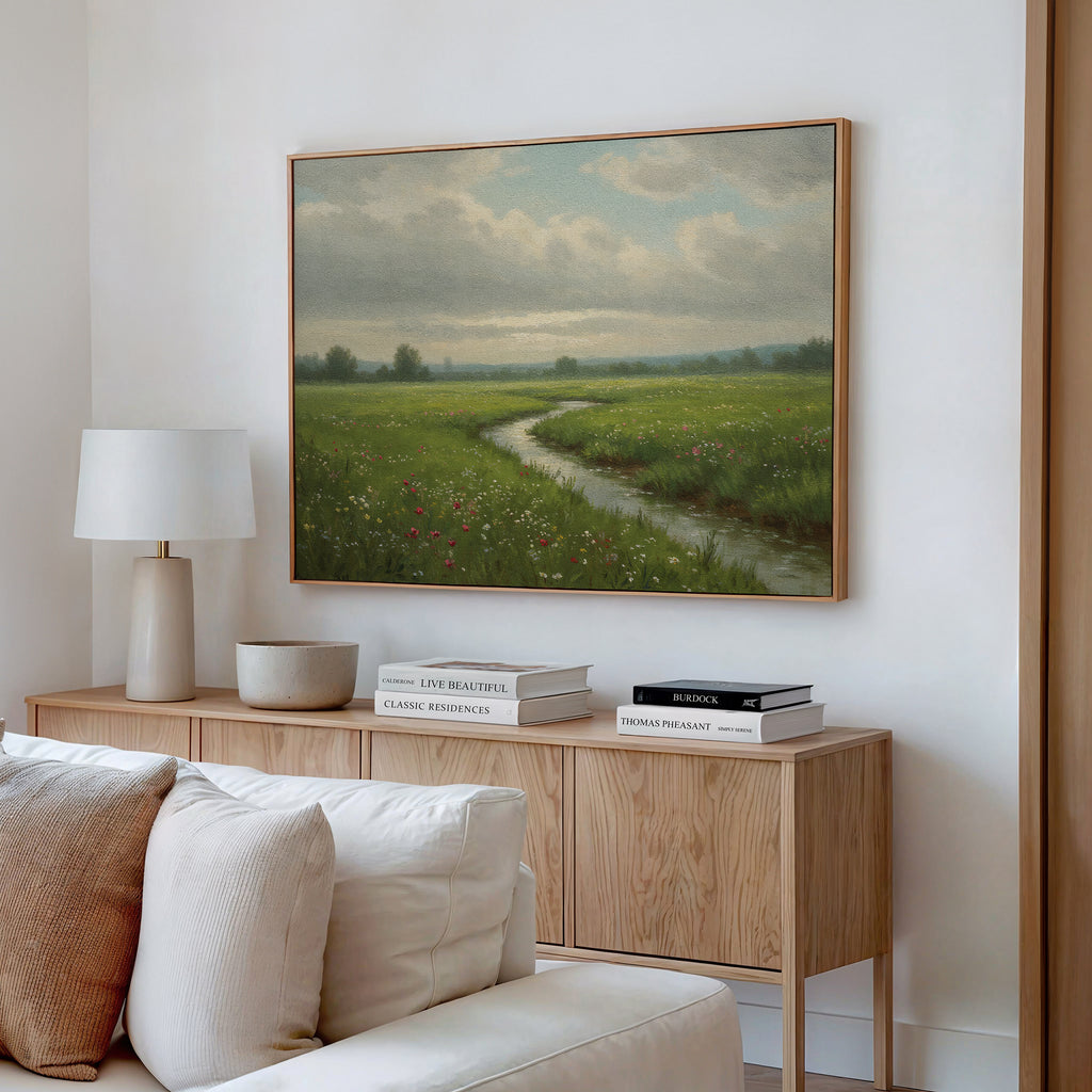 A cozy living room features a wooden sideboard with stacked books, a lamp, and a bowl. Above hangs the Winding Stream Canvas Art, depicting a grassy field and stream under cloudy skies. A beige sofa sits in the foreground.