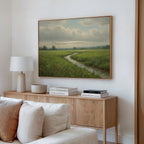 A cozy living room features a wooden sideboard with stacked books, a lamp, and a bowl. Above hangs the Winding Stream Canvas Art, depicting a grassy field and stream under cloudy skies. A beige sofa sits in the foreground.