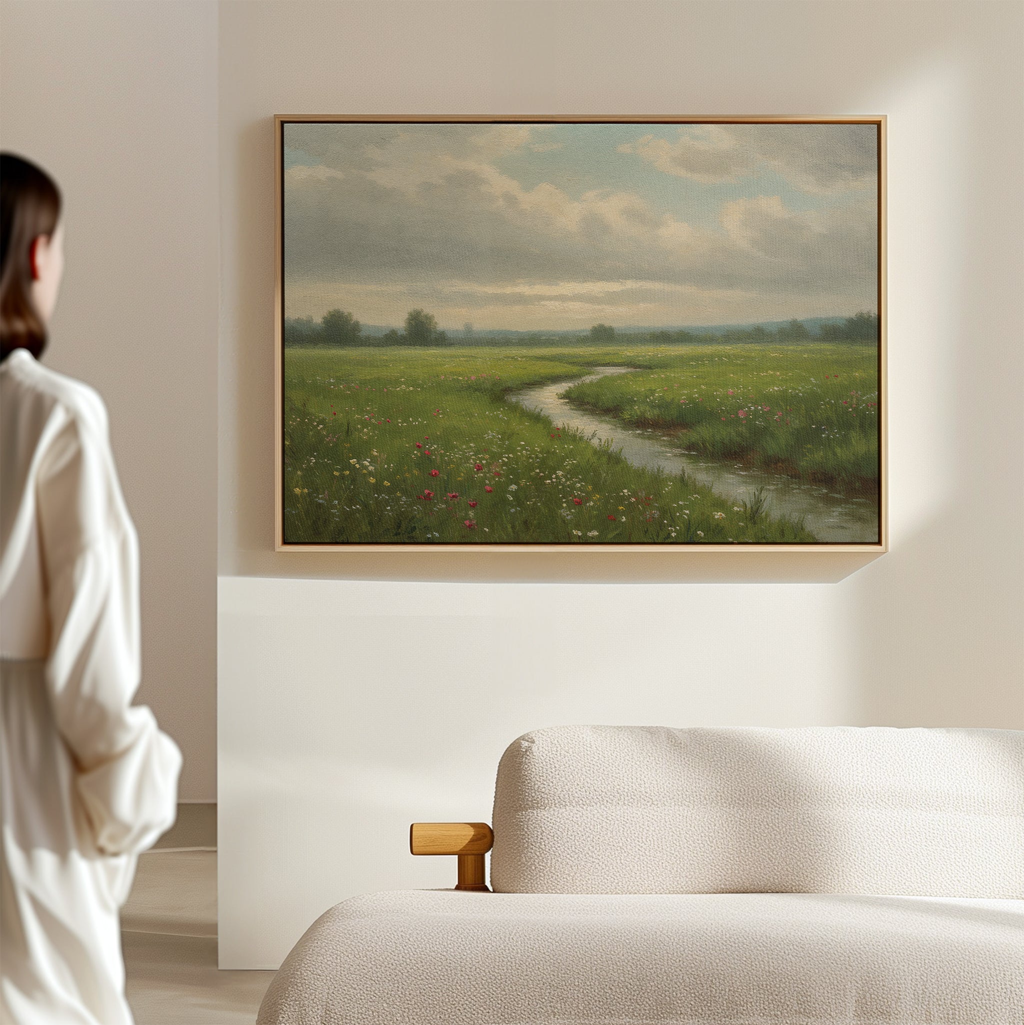 A woman admires the Winding Stream Canvas Art in a minimalist room, featuring a stream winding through a green meadow with wildflowers under a cloudy sky. A light-colored sofa in the foreground enhances the serene display.