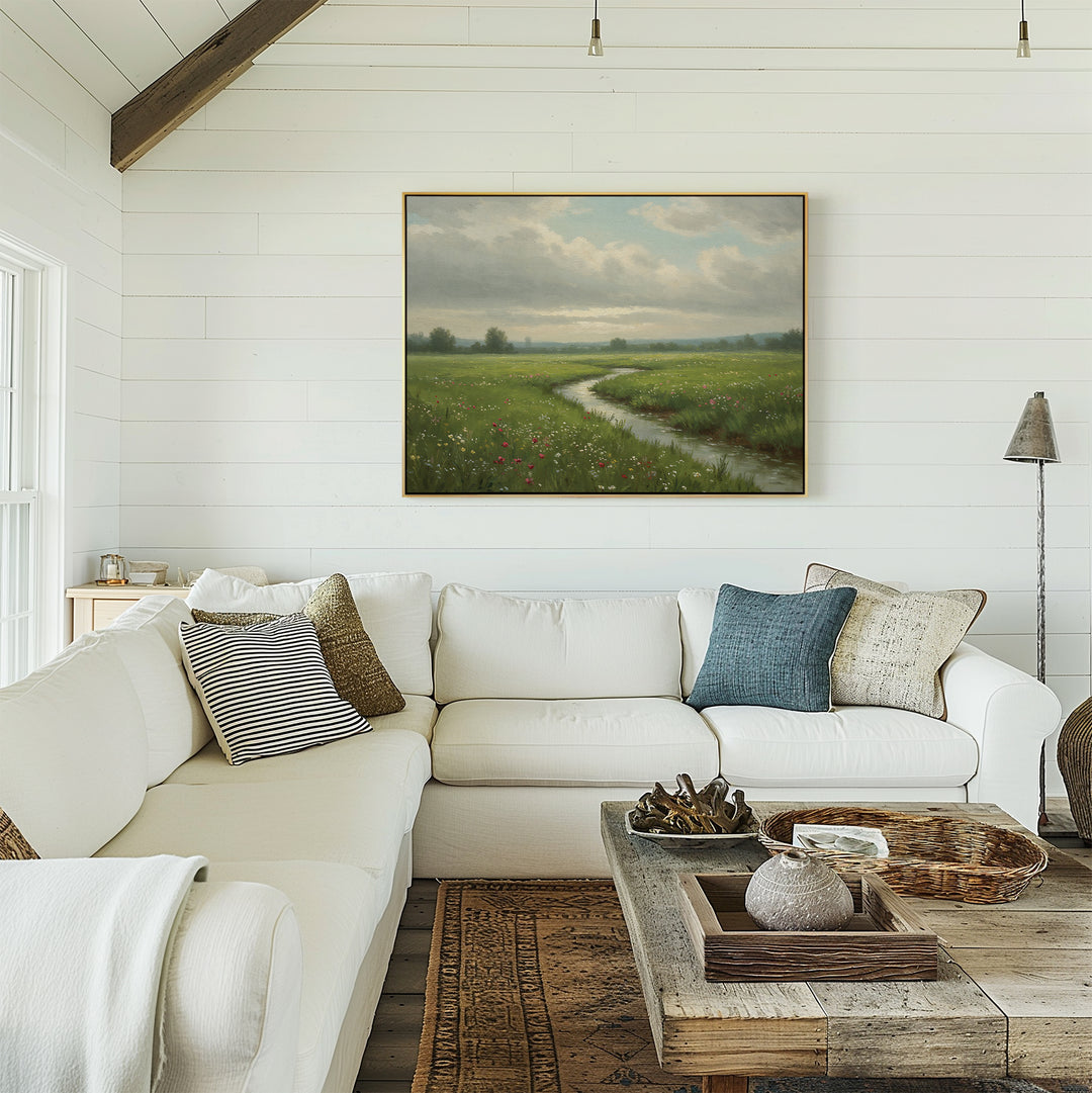A cozy living room with a white sectional sofa, assorted pillows, a rustic wooden coffee table, and the Winding Stream Canvas Art—a large green meadow landscape—displayed on the shiplap wall.