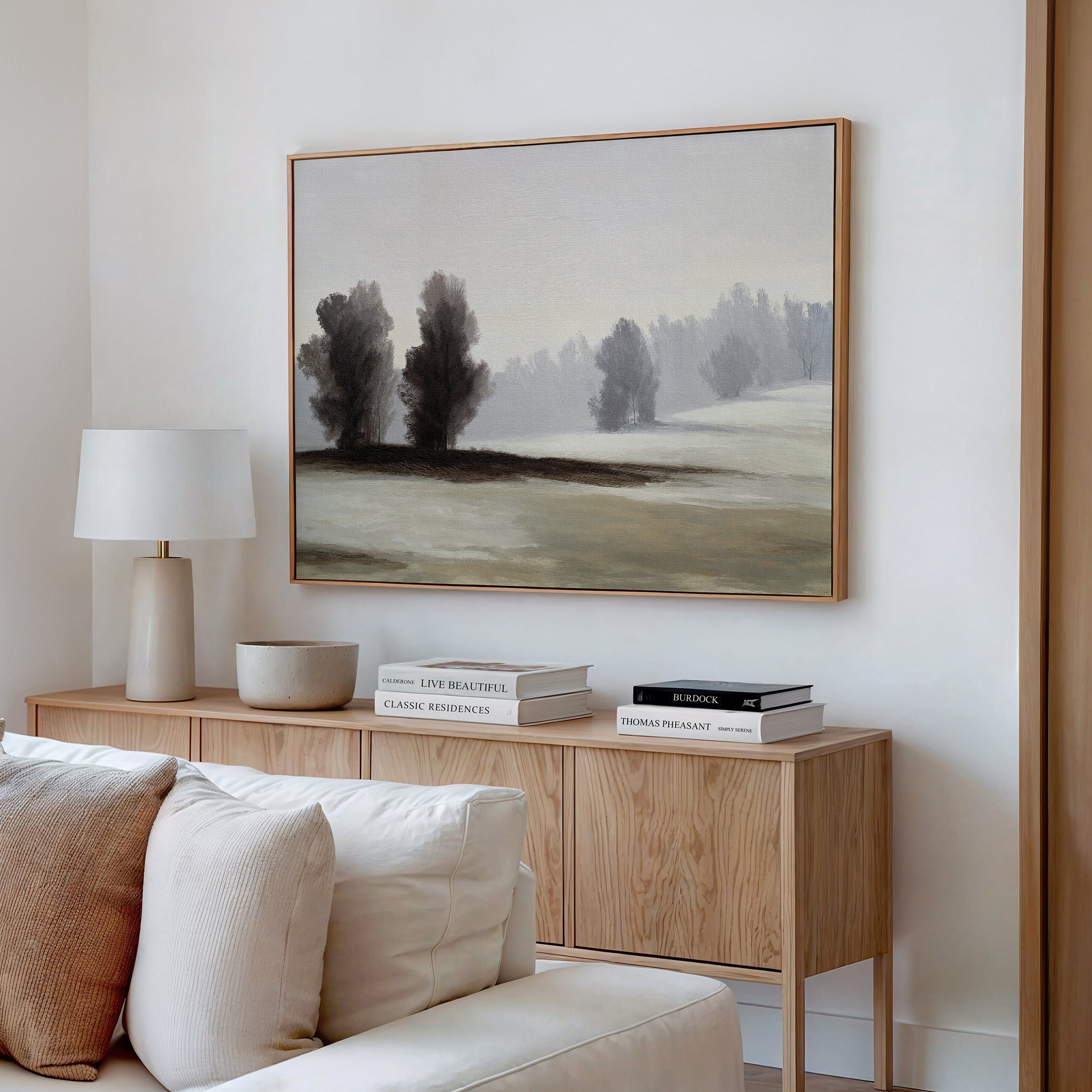 A minimalist living room features a light wood sideboard, neutral decor, white lamp, stacked books, and the Winter Dusk Canvas Art—showcasing serene trees and hills—framed above a beige sofa with cozy cushions.