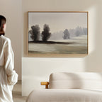 A person in white stands in a bright, minimal room, admiring Winter Dusk Canvas Art—a minimalist artwork of trees and fields—displayed above a beige textured sofa.